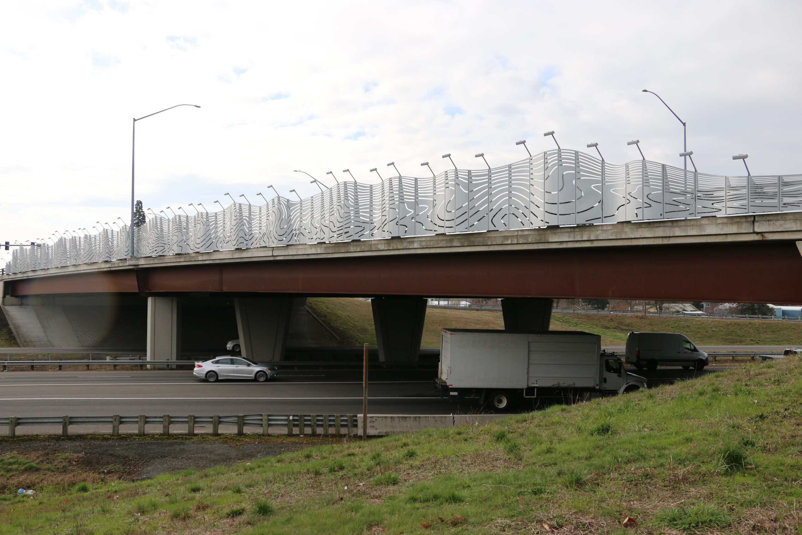 A large silver wavy sculpture that lives above a highway with cars passing by.