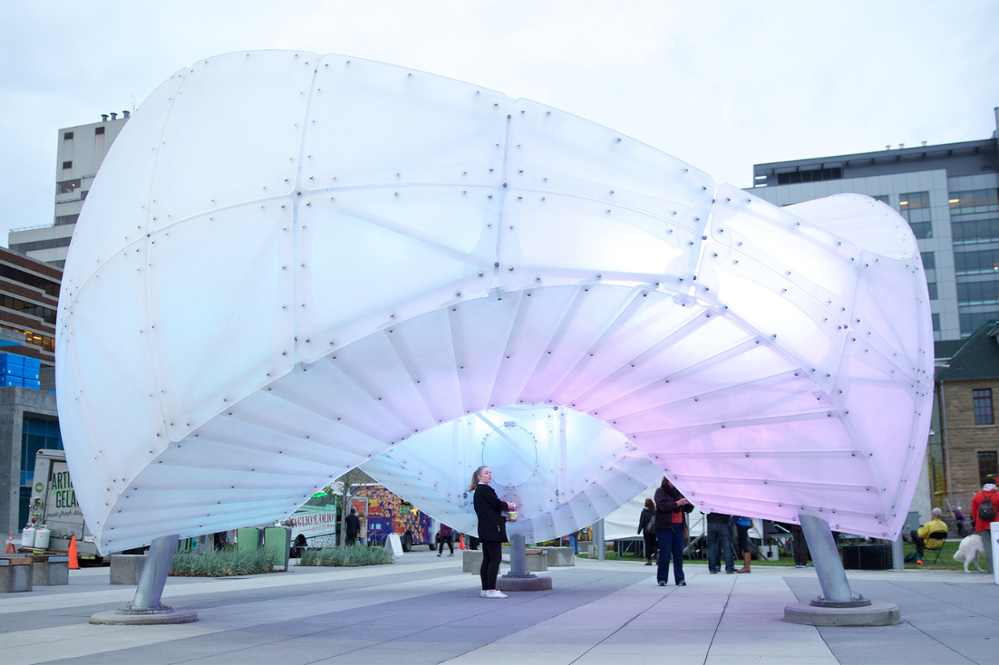 White cloud-like shape sculpture that is hovering over the ground where people can stand underneath in an open park.