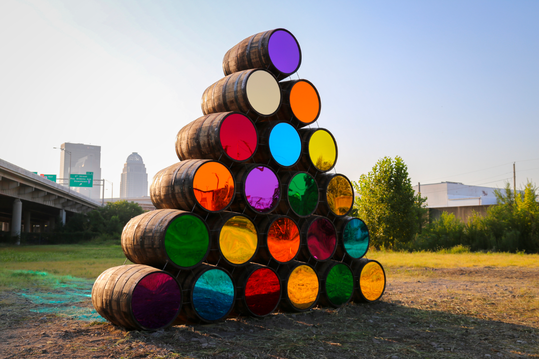 Twenty colorful barrels are placed on top of one another, forming a pyramid outside in the city.